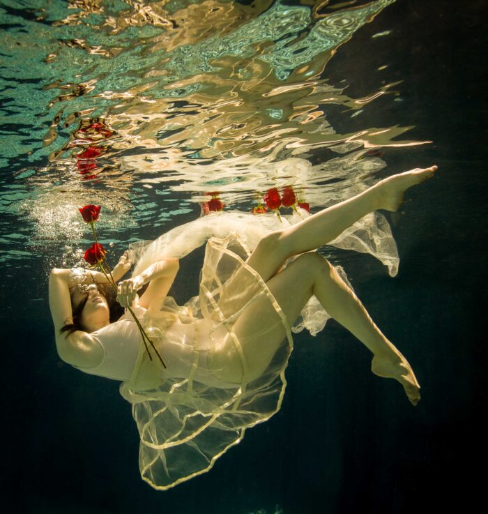 Underwater photograph of a figure in a pale gown holding red roses