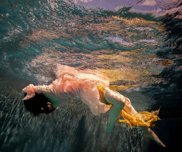 Underwater photograph of a figure in a pale pink dress with gold fabric trailing in the current